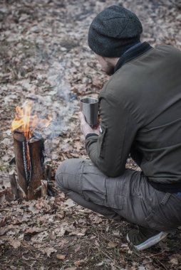 Bir adam demir bardaktan çay içer ve ateşe bakar.,