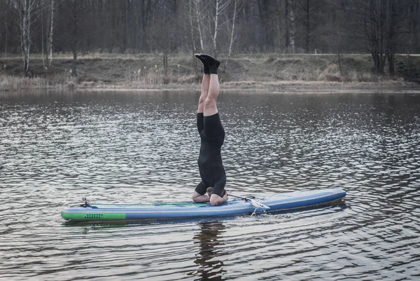 Sup Yoga, bulutlu bir günde yoga yapan dalgıç kıyafetli bir adam, kürek tahtasında amuda kalkıyor., — Stok fotoğraf