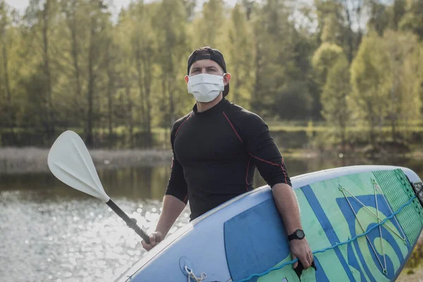 Homem atlético em uma máscara médica fica com uma pá no fundo do lago, atleta paddleboard , — Fotografia de Stock
