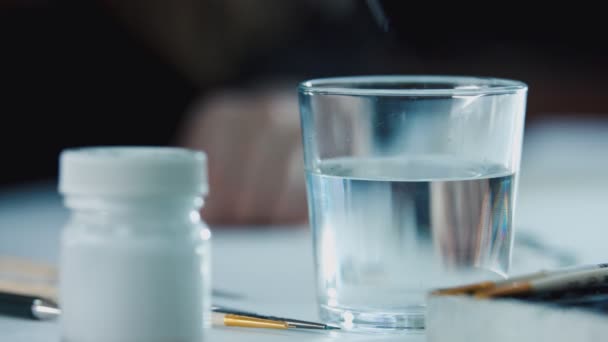 Frau reinigt Pinsel von blauer Aquarellfarbe in einem Glas sauberen Wassers und macht abstrakte Linien auf Wasseroberfläche — Stockvideo