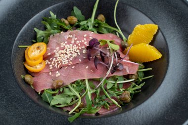 Kapari, roka ve siyah tabakta portakal kabuğuyla parma jambonu salatası yakın.