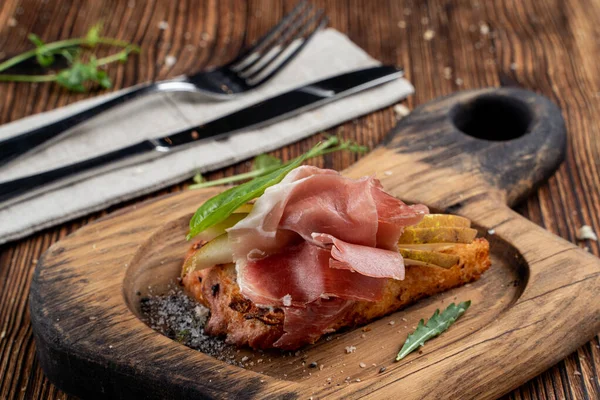Parma jambonlu bruschetta, yumuşak peynirli ve olgun armutlu tost, ahşap kesim tahtasında servis ediliyor.
