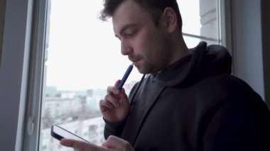 Şaşırmış genç adam telefonunu kontrol ediyor, camda sigara içiyor, kapatıyor.