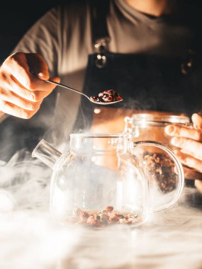 Barista, hafif dumanla çevrili bir barın dramatik ışığında kuru gül goncalarıyla gül çayı, cam çaydanlık dolduruyor; yakın çekim, eller görünür