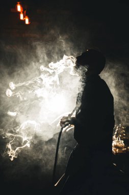 Genç bir adamın silueti, nargile tüttürüyor ve koyu arkaplanda yoğun dumanlar salıyor.