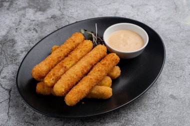 Bol yağda kızartılmış mozzarella peynirli çubuklar içinde erimiş peynir, siyah tabakta sosla servis edilmiş, minimalist atış.