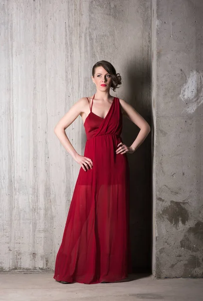Retrato vertical de uma bela jovem mulher em vestido vermelho longo em pleno crescimento em um fundo de paredes de concreto cinza — Fotografia de Stock