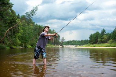 Fishing before the storm. A young man catches a fish on spinning, standing in the river clipart