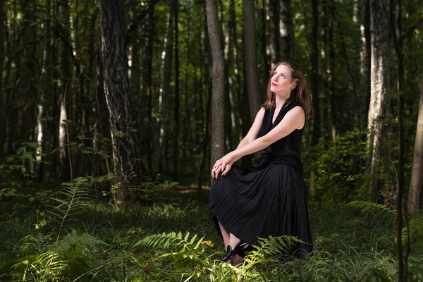 Delícia na natureza. Retrato de uma jovem mulher em um vestido preto sentado na floresta — Fotografia de Stock
