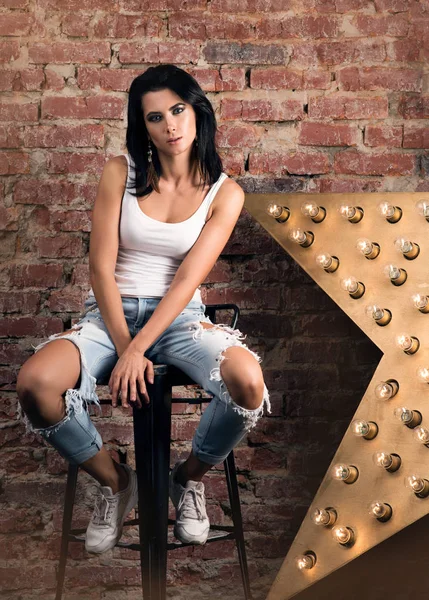 Modern young woman posing on a chair. Stylish appearance, white  t-shirt and jeans with holes