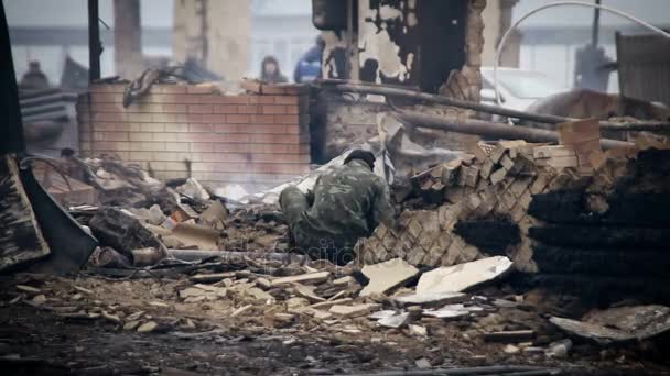 Ein junger Mann in Tarnkleidung gräbt in den verbrannten Überresten des Hauses. Blick auf das abgebrannte Dorf — Stockvideo