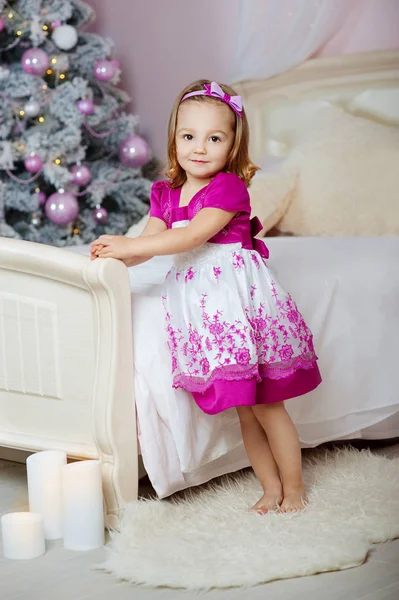 Mignonne petite fille dans une robe rose debout près du lit sur le fond de l'arbre de Noël — Photo