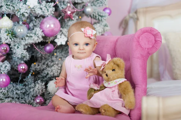 Charmante petite fille aux yeux bleus assise dans un fauteuil rose ours en peluche sur fond décoré arbre de Noël — Photo