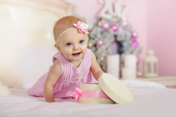 Petite fille dans une robe rose avec des fleurs dans ses cheveux sourit et ouvre la boîte cadeaux sur le lit le fond des décorations de Noël — Photo