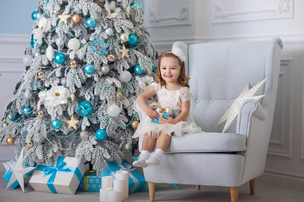 cute little girl in bklom dress sitting in a chair and opens box with present for background Christmas tree blue ornaments
