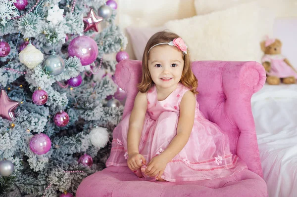 Belle petite fille assise et pose sur une chaise. arrière-plan festif sapin ensemble de feux lumineux. Souriez doucement. Noël concept de vacances — Photo