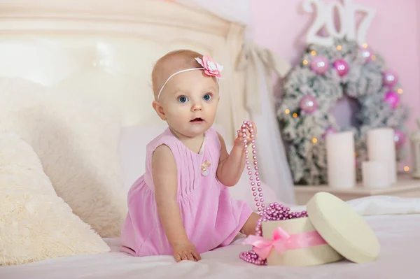 Petite fille dans une robe rose avec des fleurs dans ses cheveux sourit et ouvre la boîte cadeaux sur le lit le fond des décorations de Noël — Photo
