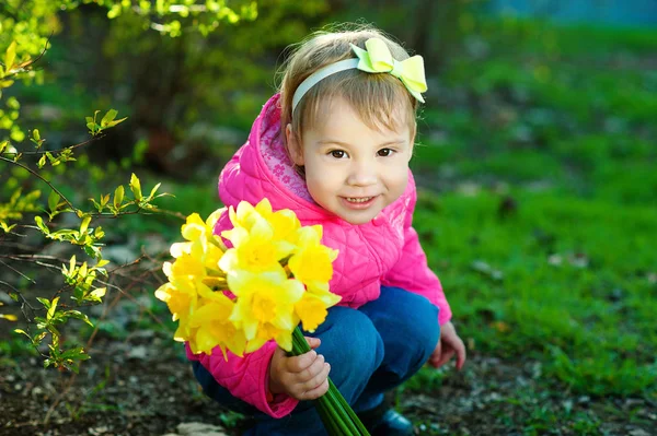 Menina Com um arco no cabelo sentado na grama e segurando um buquê de narcisos amarelos. Uma criança caminha para fora em tempo quente . — Fotografia de Stock