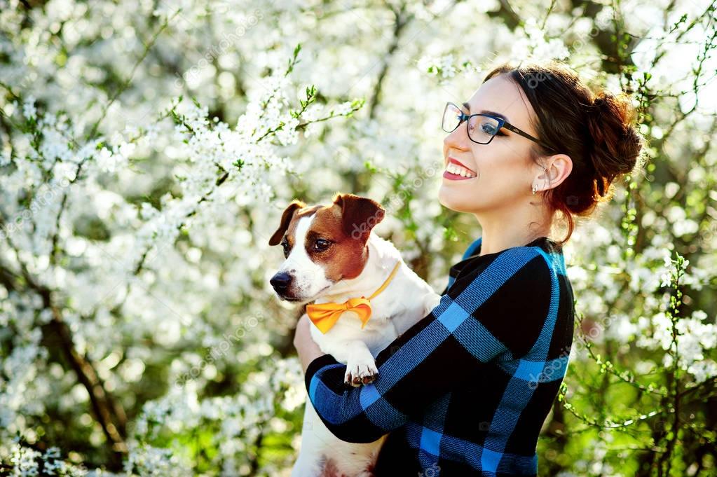 Young beautiful woman smiling and holding a dog Jack Russell terrier in the hands on a background of spring blooming trees