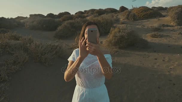 Girl in white dress makes the photo on the phone on a sunset, in desert. Warm summer evening, desert. Slow motion — Stock Video