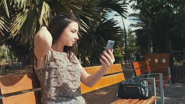 Ragazza guarda nello smartphone come uno specchio sulla panchina e toccando i capelli di fronte alla fotocamera sullo sfondo. Giovane signora che tiene smartphone di fronte a lei . — Video Stock