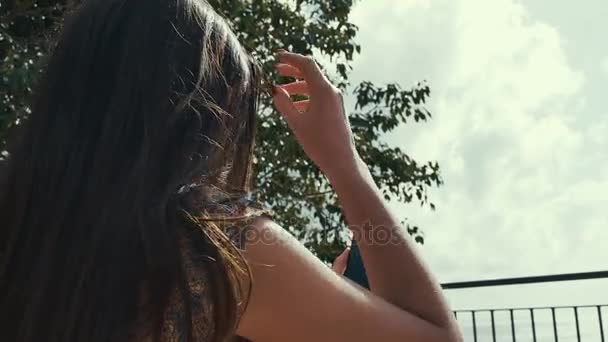 Girl looks in the smartphone as a mirror on the bench and touching her hair facing the camera on background. Young lady holding smartphone in front of her. — Stock Video