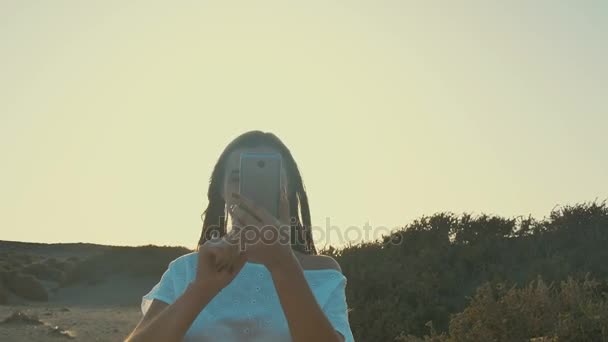 Meisje in witte jurk maakt de foto op de telefoon op een zonsondergang, in de woestijn. Warme zomeravond, ochtend, silhouet van het meisje tegen de hemel. — Stockvideo