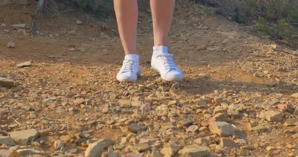 Hermosas piernas de mujer en zapatillas blancas, que van a lo largo del suelo pedregoso. Caminando en el desierto . — Vídeos de Stock