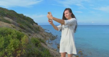 Genç güzel kadın selfie fotoğraf deniz üzerine onun smartphone kullanarak yapıyor. Gün batımı sırasında kadın ayakta deniz kenarında. Açık mavi su.