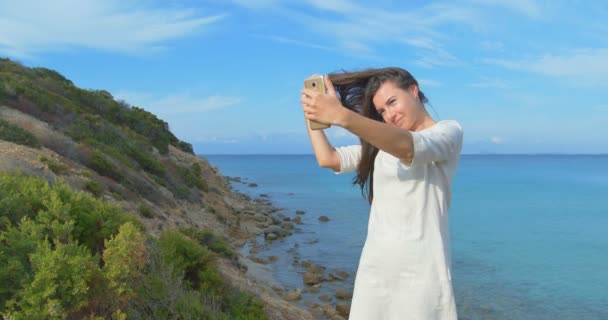 Fiatal, csinos nő selfie fénykép használ a smartphone, tengerre néző csinál. Női állt közel a tenger, naplemente alatt. Tiszta kék víz. — Stock videók
