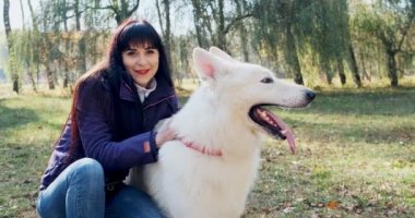 Mutlu bayan güler yüzlü bir şekilde sonbahar parkında köpekle oynuyor ve onunla oturuyor. Evcil hayvanlarla sevgi ve arkadaşlık. Beyaz Çoban