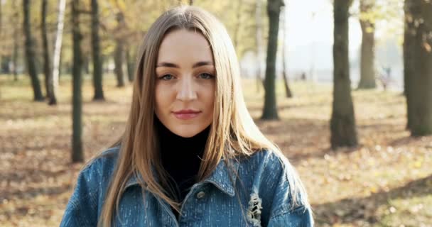 Egy fiatal, vonzó nő portréja, aki mosolyog és a kamerába néz az őszi parkban. Vidáman csinos nő a napos időben. Közelről. — Stock videók