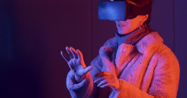 Hermosa mujer joven usando gafas de realidad virtual sobre fondo de iluminación de neón. Chica atractiva tocando objetos virtuales en gafas vr — Vídeos de Stock