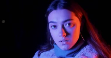 Geceleri kırmızı ve mavi ışıklı güzel esmer kadın hareket eden kameraya bakar. Makyajlı ve renkli küpeli manken kız.