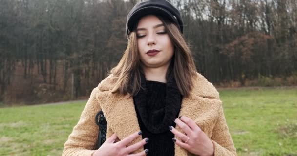 Retrato de jovem fêmea atraente que sorrindo e olhando para a câmera no gramado verde. Mulher alegremente bonita esconde seu casaco do frio. Fechar — Vídeo de Stock