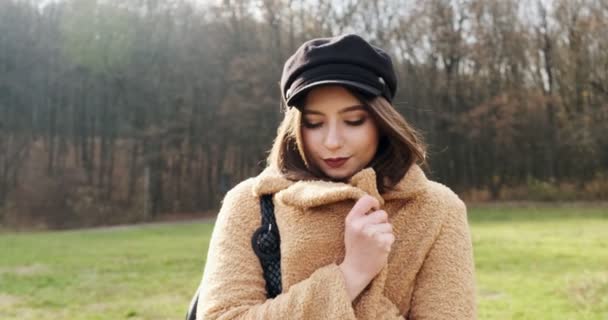 Portret młodej atrakcyjnej kobiety, która uśmiecha się i patrzy w kamerę na zielonym trawniku. Śliczna kobieta chowa się w płaszczu przed zimnem. Zamknij się. — Wideo stockowe