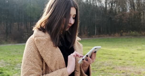 Portret młodej atrakcyjnej kobiety uśmiechającej się i patrzącej na smartfona na zielonym trawniku w jesiennym parku w pobliżu lasu — Wideo stockowe