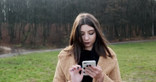 Porträt einer jungen attraktiven Frau, die auf dem grünen Rasen des Herbstparks am Wald spazieren geht und auf ihr Smartphone schaut — Stockvideo
