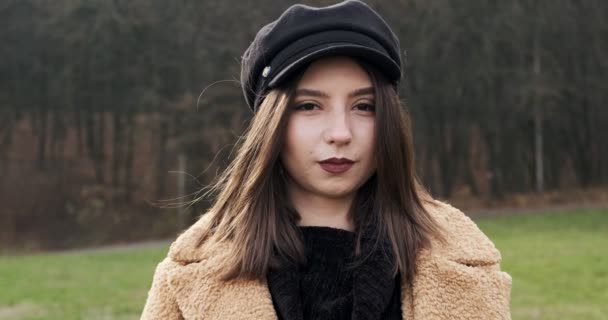 Retrato de jovem fêmea atraente que sorrindo e olhando para a câmera no gramado verde. Mulher alegremente bonita com chapéu. Fechar — Vídeo de Stock