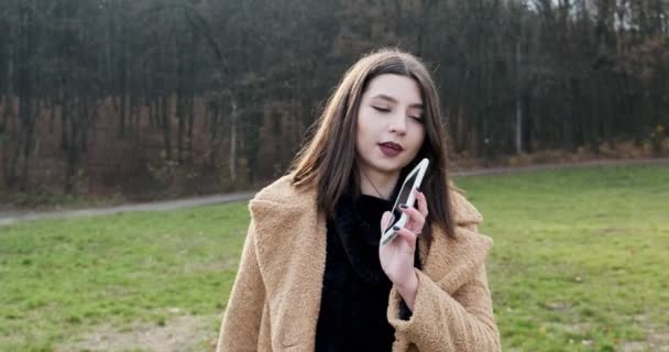 若い魅力的な女性の肖像笑顔と電話を取り上げ、森の近くの秋の公園の緑の芝生で通信します — ストック動画