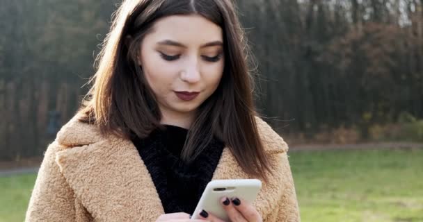 Porträtt av ung attraktiv kvinna ler och tittar på smartphone på den gröna gräsmattan i höstparken nära skogen — Stockvideo