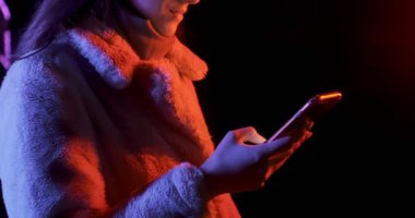 Geceleri kırmızı ve mavi neon ışıklı güzel esmer kadın akıllı telefona bakıyor. Makyajlı, renkli ışıklı ve telefonlu küpeli manken kız.
