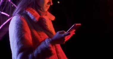 Geceleri kırmızı ve mavi neon ışıklı güzel esmer kadın akıllı telefona bakıyor. Makyajlı, renkli ışıklı ve telefonlu küpeli manken kız.