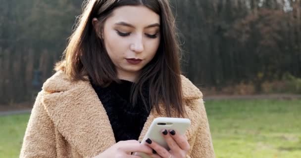 Retrato de una mujer joven y atractiva sonriendo y mirando el teléfono inteligente en el césped verde en el parque de otoño cerca del bosque — Vídeos de Stock