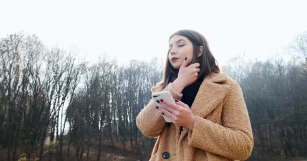 Retrato de una mujer joven y atractiva sonriendo y mirando el teléfono inteligente en el césped verde en el parque de otoño cerca del bosque — Vídeos de Stock