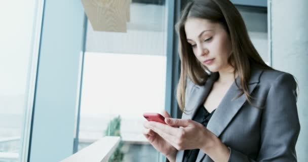 Porträt einer jungen attraktiven Frau, die im Büro in der Nähe des Fensters auf ihr Smartphone blickt — Stockvideo