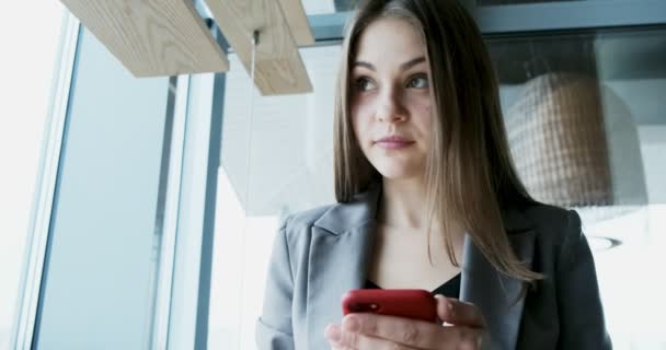 Porträt einer jungen attraktiven Frau, die im Büro in der Nähe des Fensters auf ihr Smartphone blickt — Stockvideo