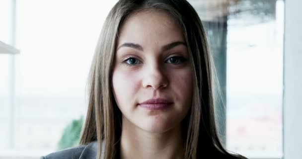 Retrato de una joven atractiva hembra que mira a la cámara en la oficina cerca de la ventana. Una mujer alegremente bonita. De cerca. — Vídeos de Stock