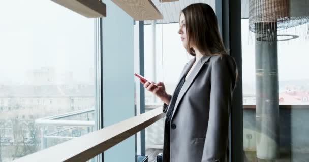 Porträt einer jungen attraktiven Frau, die im Büro in der Nähe des Fensters auf ihr Smartphone blickt — Stockvideo