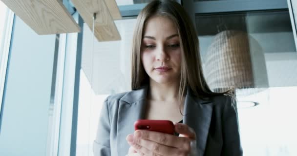 Porträt einer jungen attraktiven Frau, die im Büro in der Nähe des Fensters auf ihr Smartphone blickt — Stockvideo